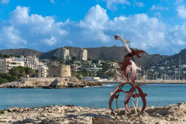 Escultura de aves metálicas en primer plano, con el castillo de Moraira y el puerto al fondo. Descubre qué ver en Moraira.