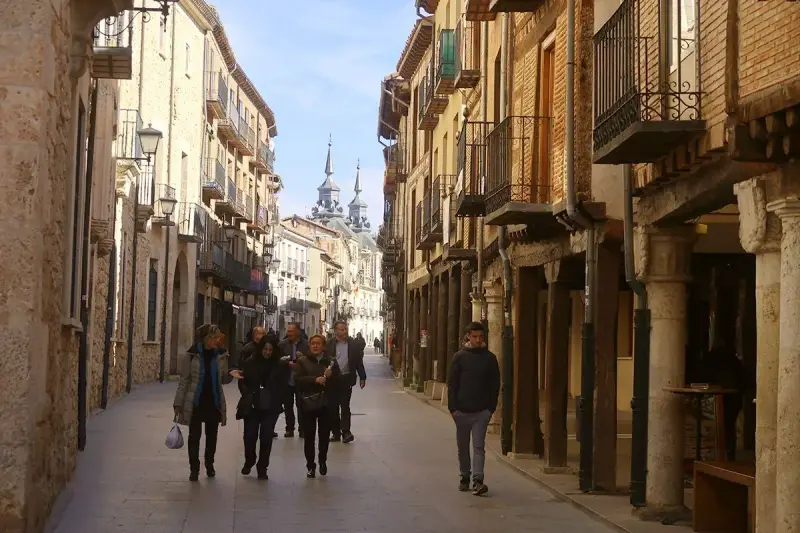 Gente pasea por una calle porticada, un lugar ideal para descubrir qué ver en Burgo de Osma. Edificios históricos y torres se alzan al fondo.