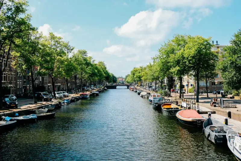 Canal de Ámsterdam con barcos, árboles y edificios bajo un cielo azul con nubes.